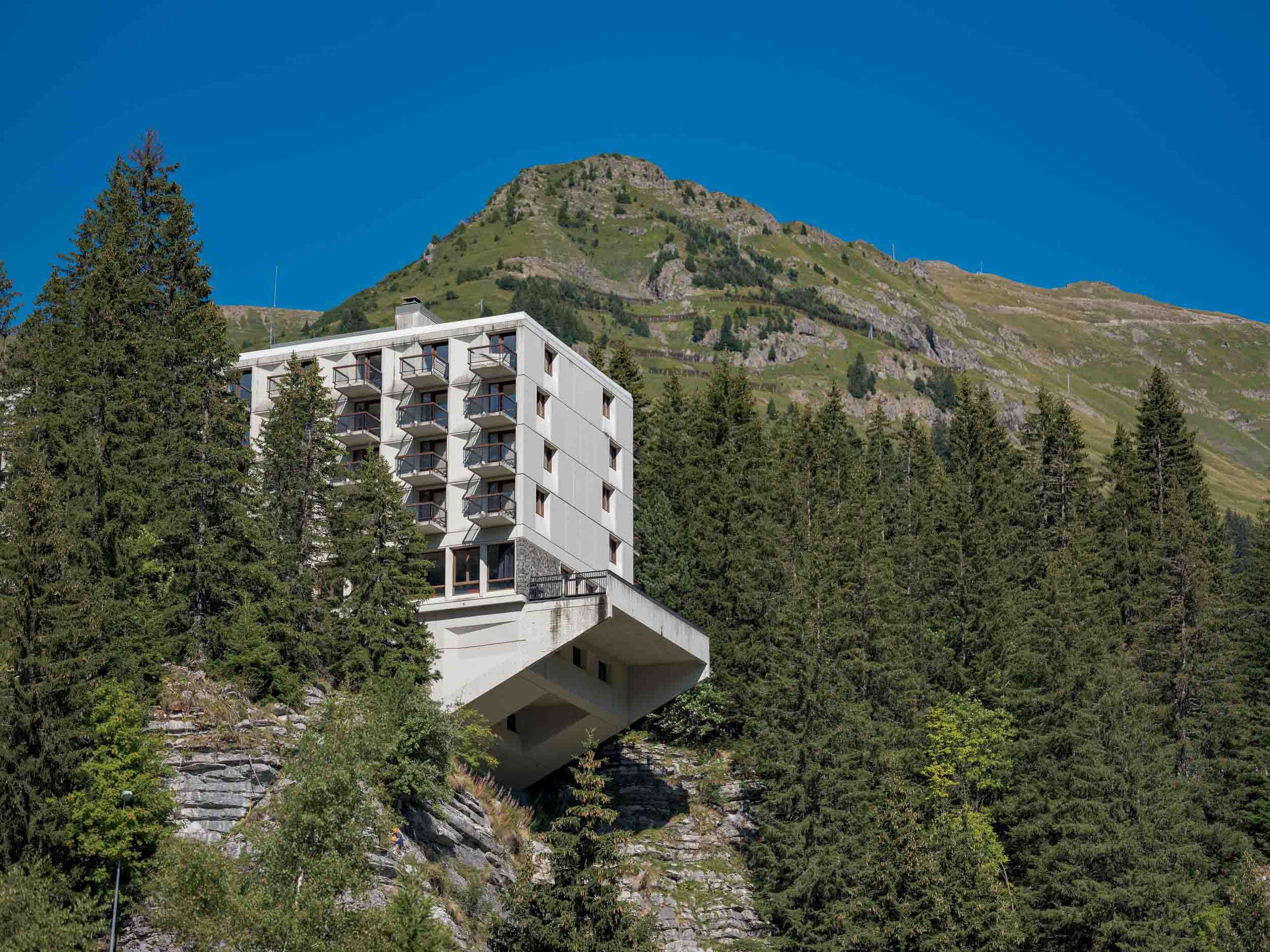 Photo du bâtiment moderne l'Hotel Le Flaine et avant-gardiste classé au l'inventaire des Monuments Historiques. à Flaine, entouré de forêts alpines avec une montagne en arrière-plan, illustrant l'architecture intégrée au paysage naturel.