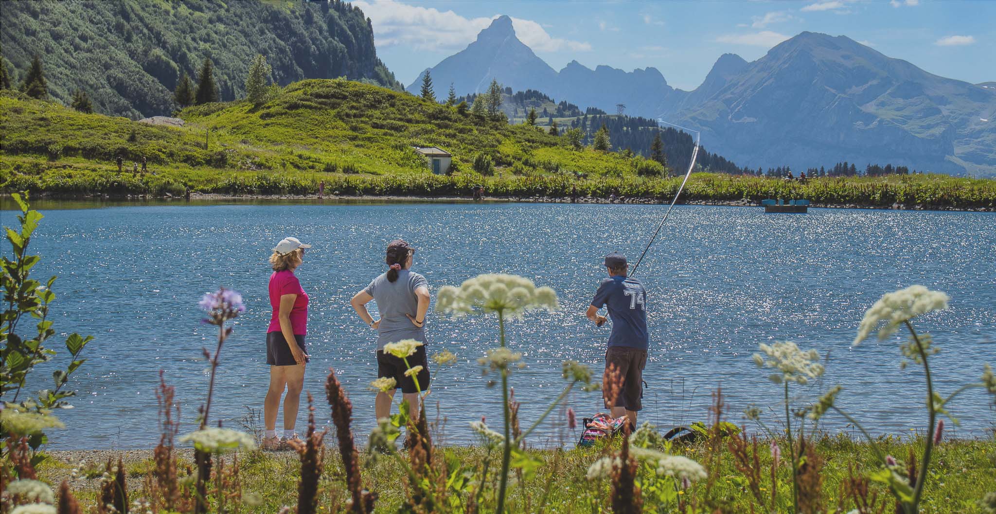 Deux touristes et un animateur de l'Office de Tourisme de Flaine participent à un atelier pêche au Lac de Vernant, avec la Pointe Percée et la chaîne des Aravis en arrière-plan.