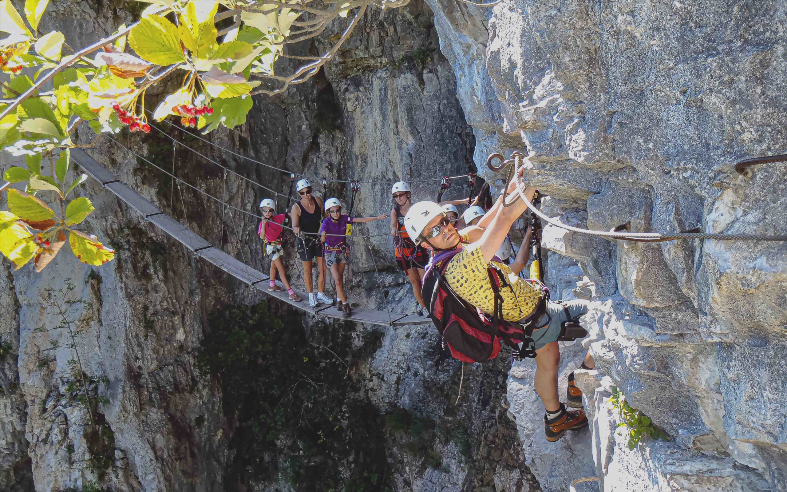 A group of holidaymakers set off on a via ferrata, crossing a rock face with safety equipment, while enjoying a spectacular view.