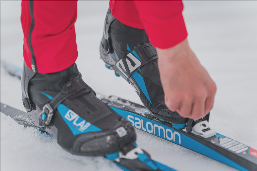 Close-up of a pair of Salomon Nordic ski boots, with a hand adjusting the binding on the skis.