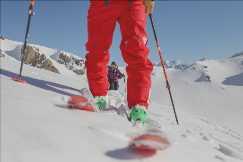 Flaine - Een vrouwelijke skitourer verschijnt tussen de benen van een mannelijke skiër op een helling vol poedersneeuw, met besneeuwde bergtoppen op de achtergrond.