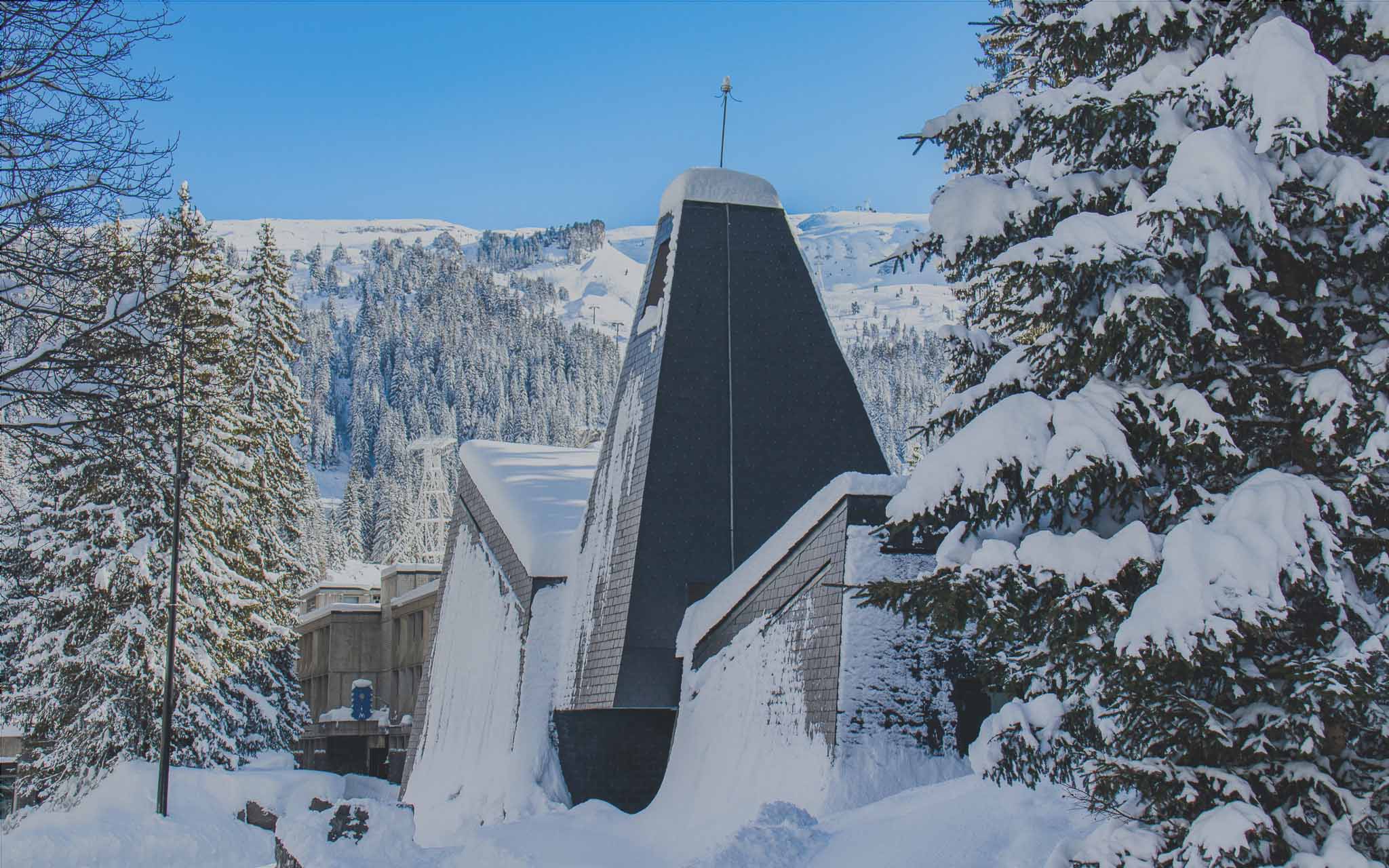 De Oecumenische Kapel in Flaine omringd door sneeuw en dennenbomen, met de bergen op de achtergrond.