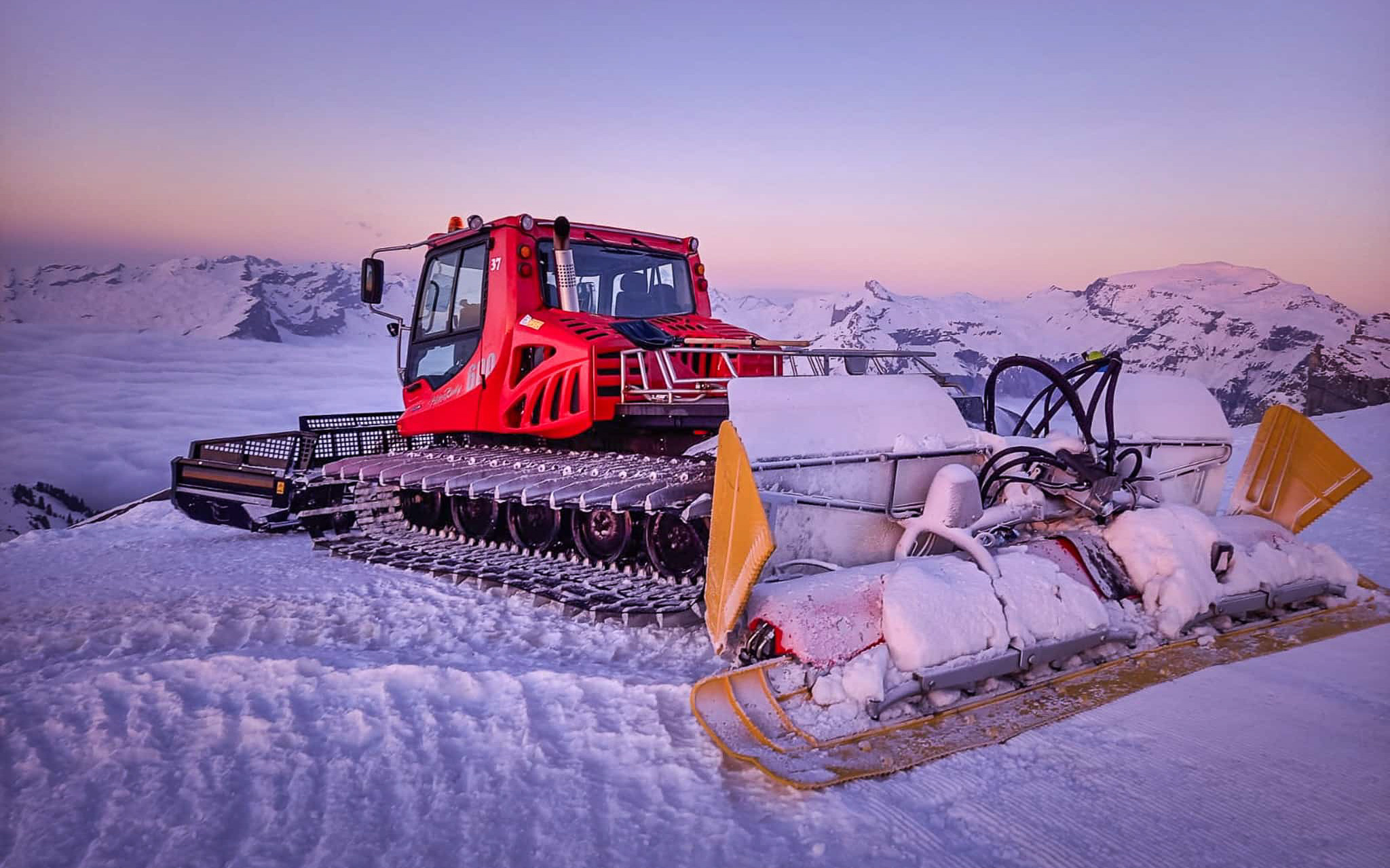 Red groomer preparing the snow-covered ski slopes of Flaine at sunset, with a mountainous landscape in the background.