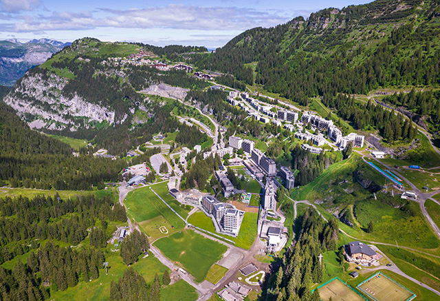 Aerial view of the resort of Flaine in summer, with its modern buildings set against a natural backdrop of mountains and lush green forests.