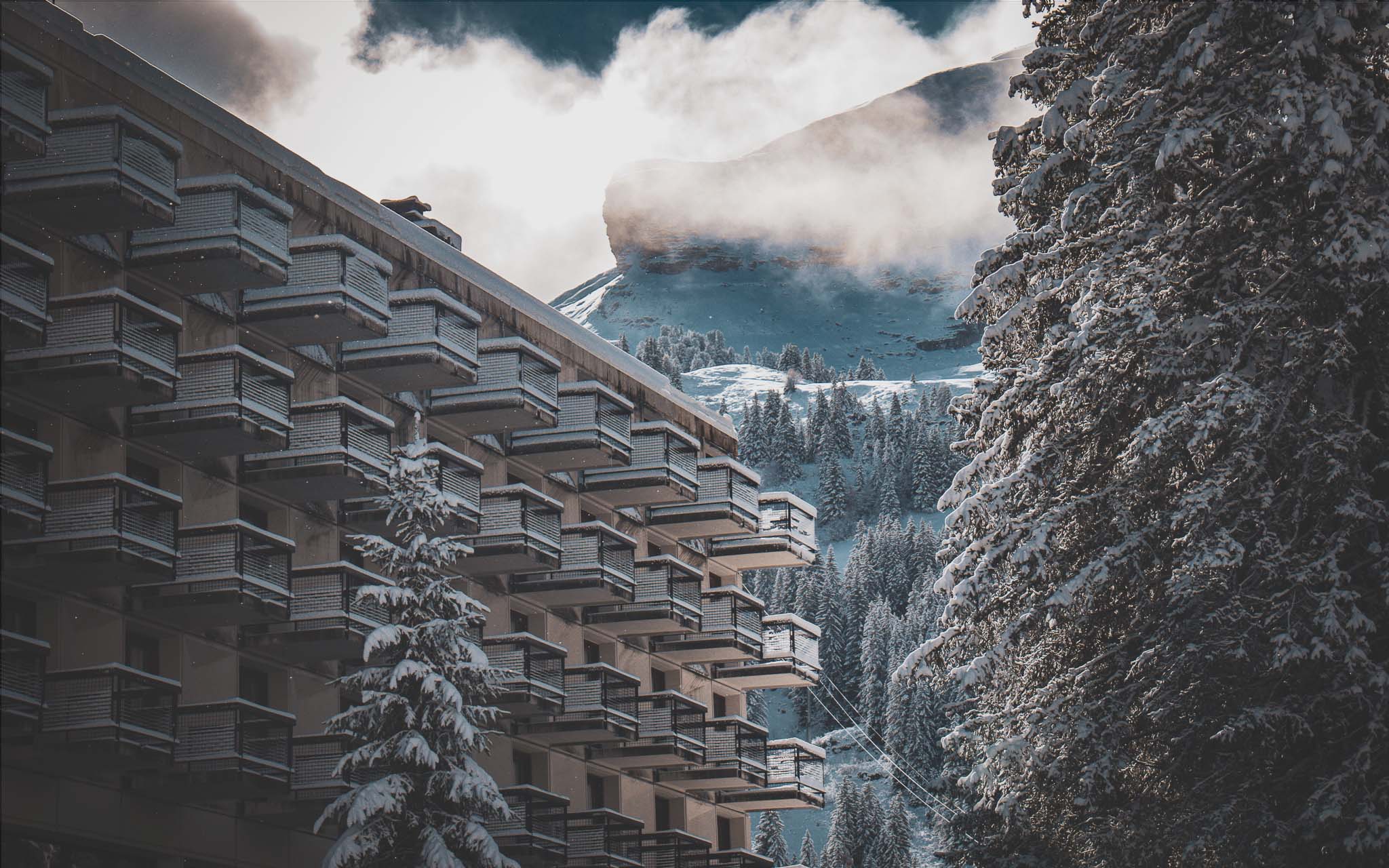 Zicht op de besneeuwde gevel van een gebouw in Flaine, met besneeuwde dennenbomen en bergen op de achtergrond