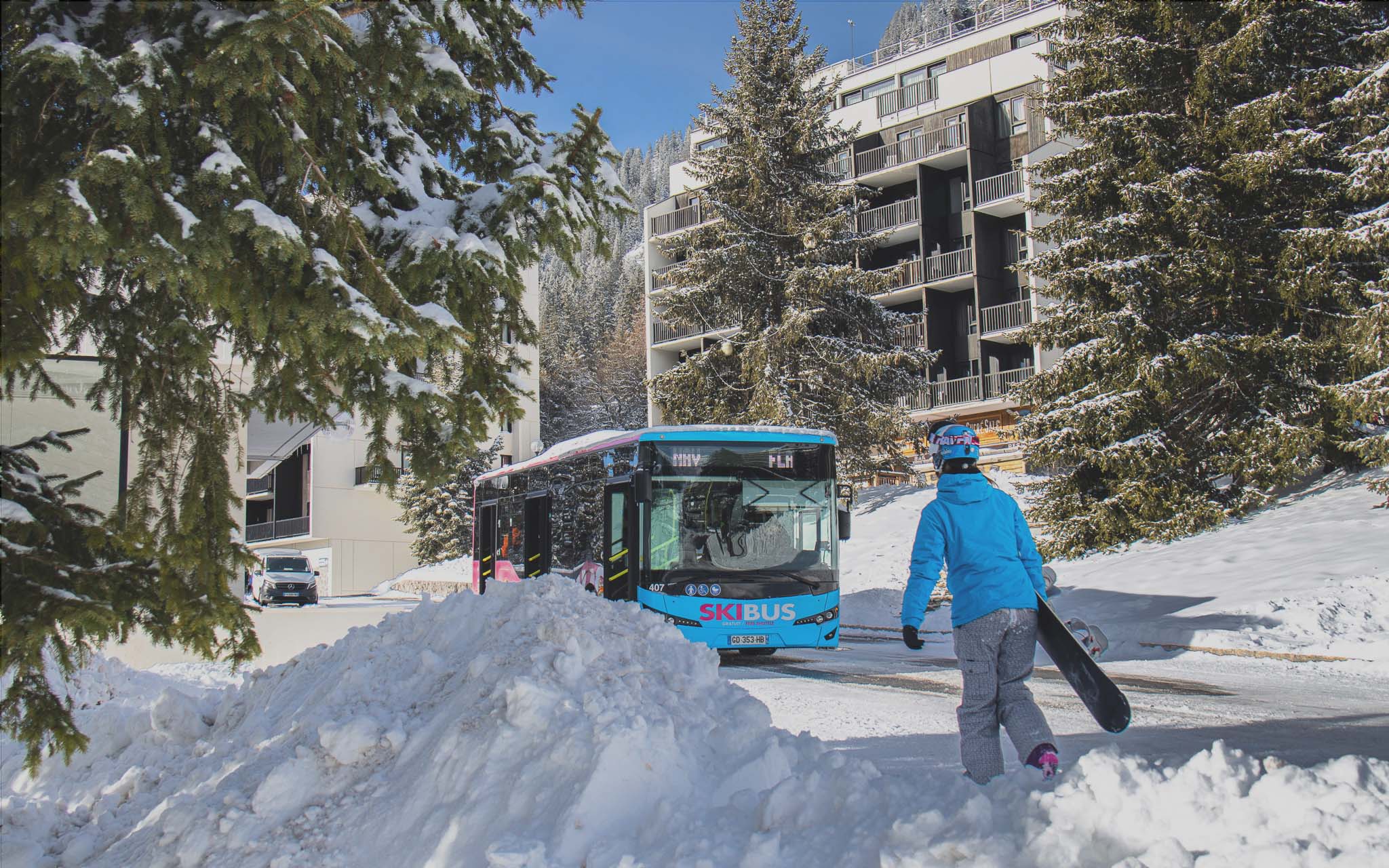 Flaine- Een snowboarder loopt naar een gratis blauwe pendelbus in het besneeuwde skigebied.