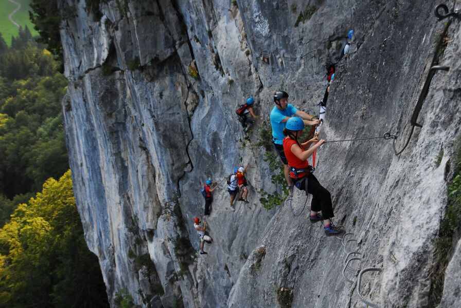 People climbing a rock face on the Via Ferrata des Monts in Sixt Fer à Cheval, using cables and ladders attached to the cliff.