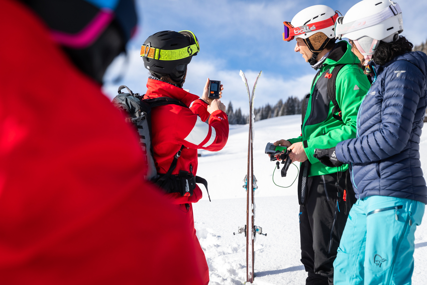 A guide in red overalls demonstrates the use of an avalanche transceiver to two skiers in the mountains, during a ski touring session on snow.