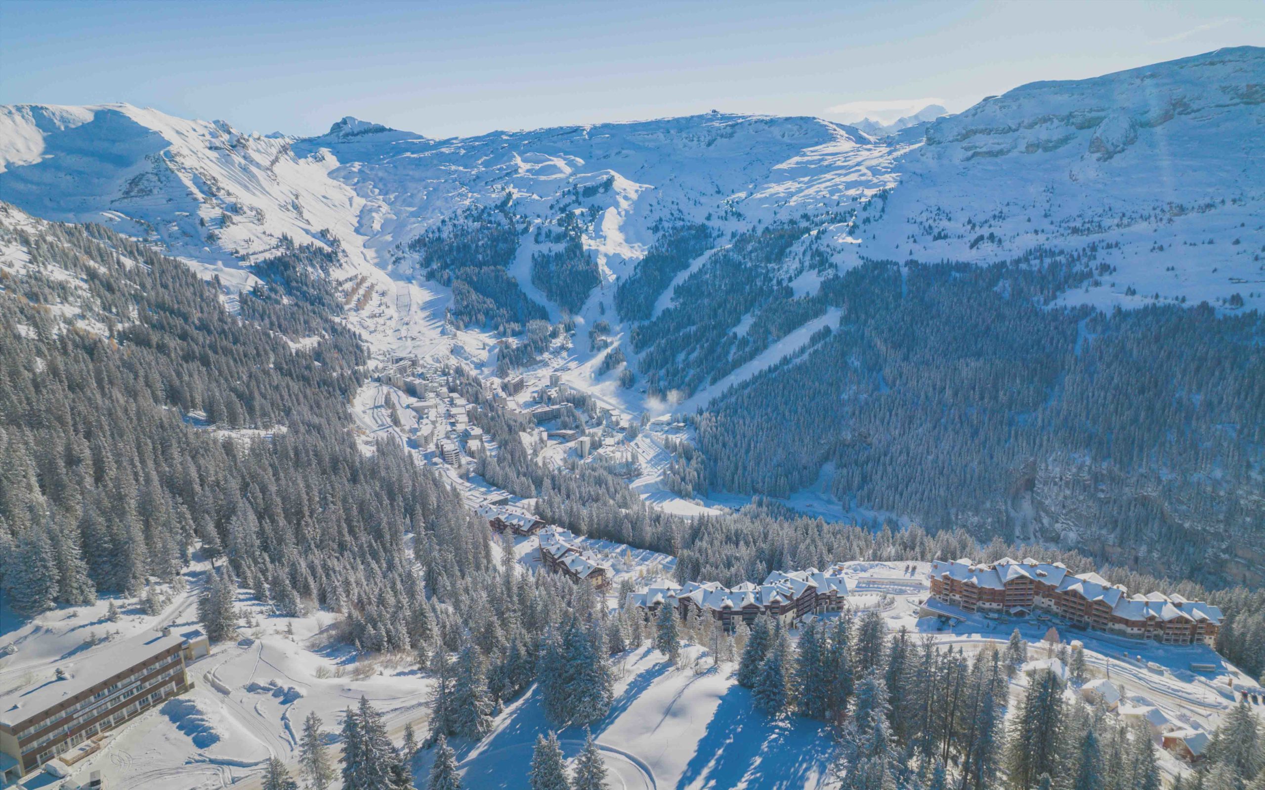 Image aérienne de la station de ski de Flaine, montrant les pistes de ski, les résidences et les forêts alpines sous un ciel bleu clair.