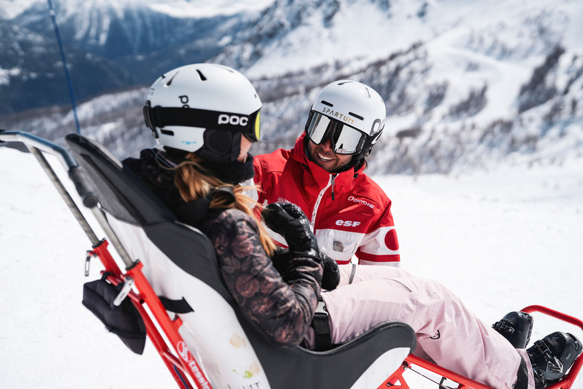 Moniteur ESF en tenue rouge aidant une skieuse en fauteuil-ski sur une piste de montagne enneigée à Flaine.