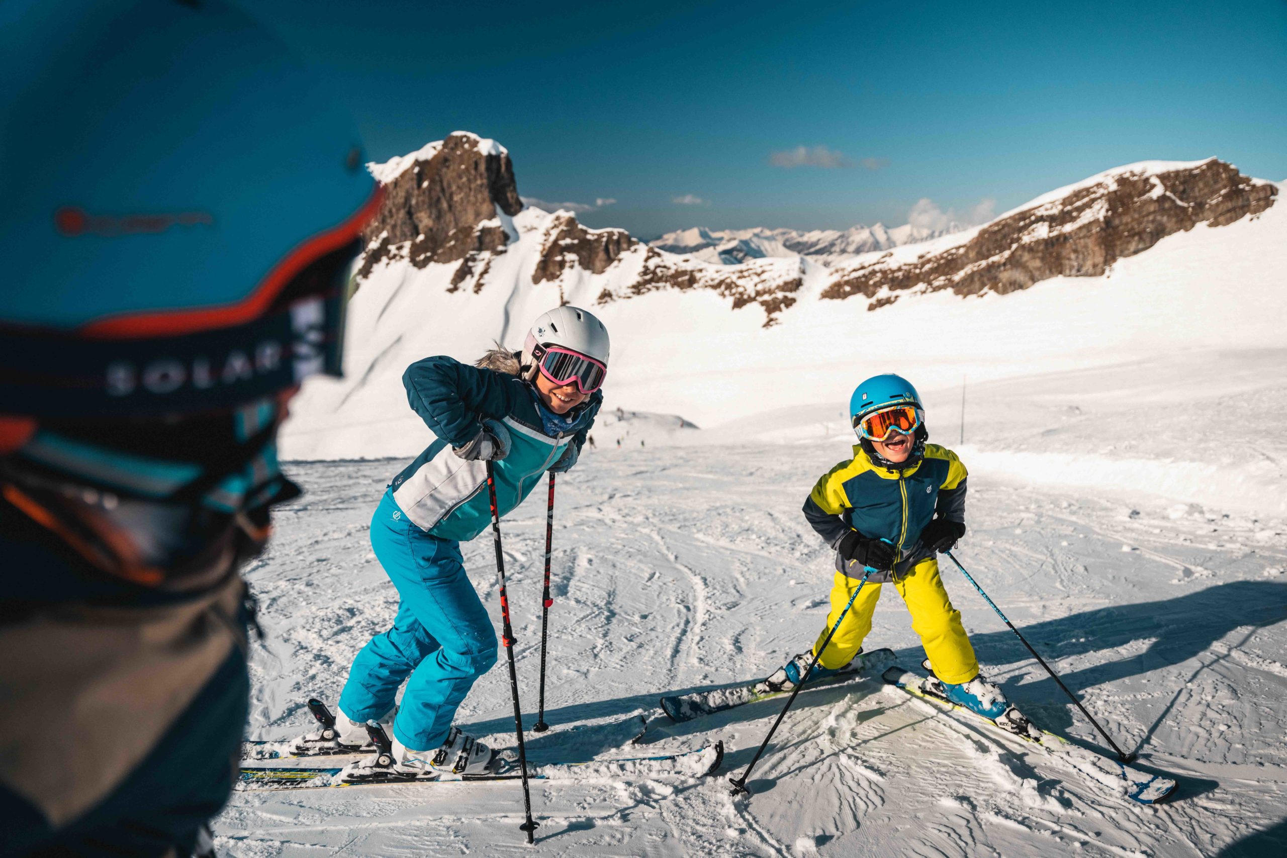 Smiling children learning to ski on the snowy slopes of Flaine, accompanied by an adult, under a sunny sky.