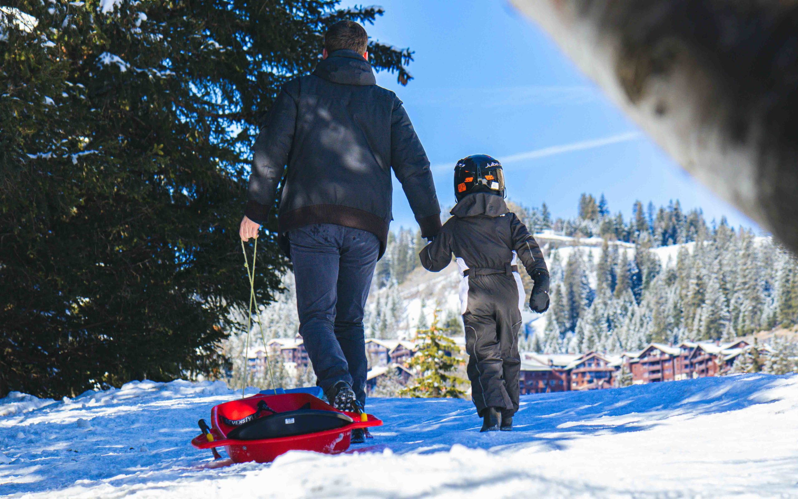 Een ouder en kind trekken een rode slee en lopen hand in hand door de sneeuw, in de besneeuwde omgeving van Flaine.